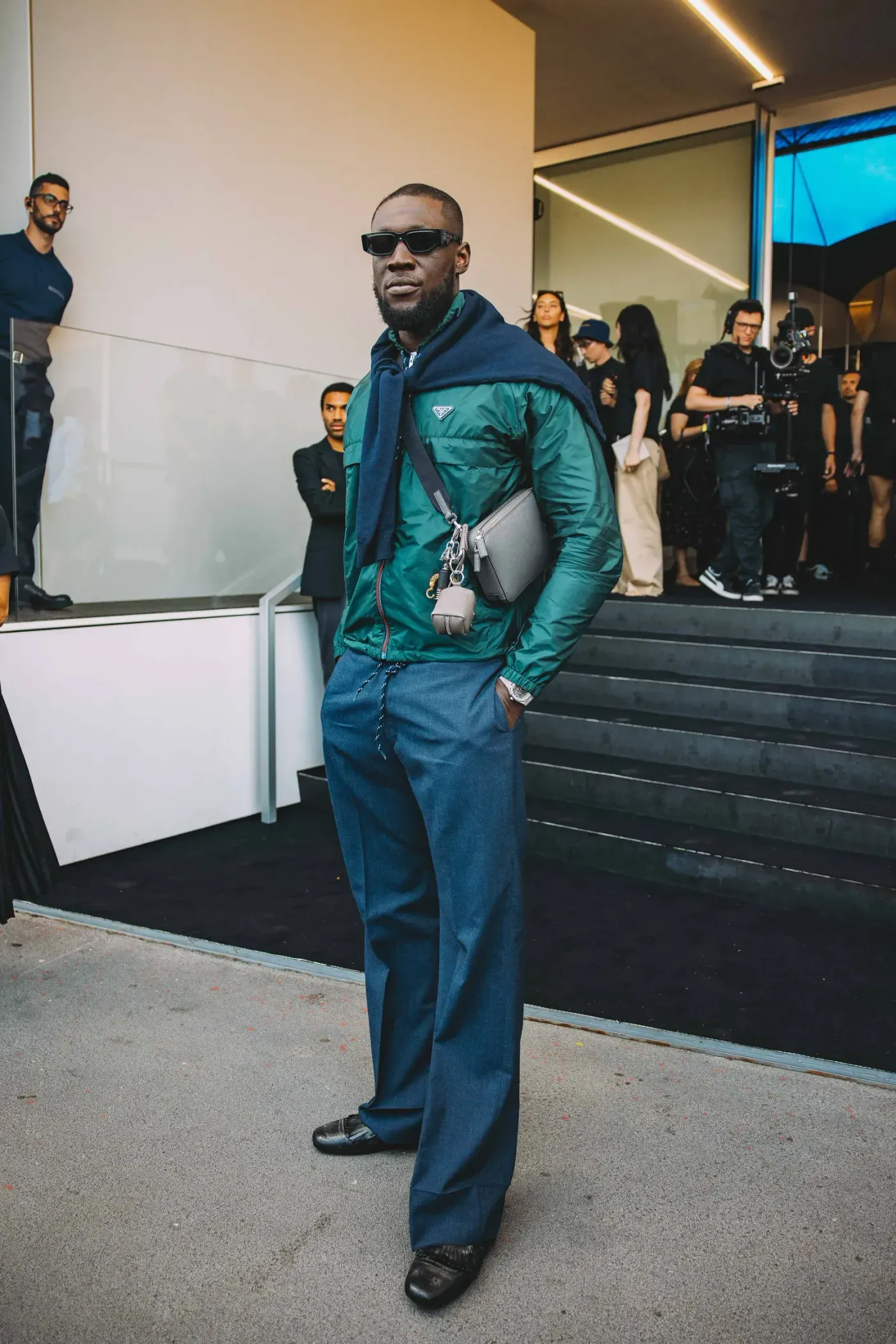 Imagen que muestra a Stormzy de pie, vestido con ropa elegante, sosteniendo una bolsa y con accesorios, en un evento de moda.