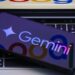 A phone displaying the Gemini logo lays on a keyboard of a laptop that has the Google logo on it.