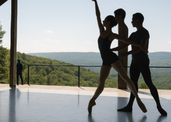 Four female dancers in white leotards, white tights, and white slippers do an arabesque facing stage left in a staggered line. They bend their supporting knee, pressing over their standing foot, and hodl their arms up in a V shape, fists clenched.