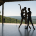 Four female dancers in white leotards, white tights, and white slippers do an arabesque facing stage left in a staggered line. They bend their supporting knee, pressing over their standing foot, and hodl their arms up in a V shape, fists clenched.